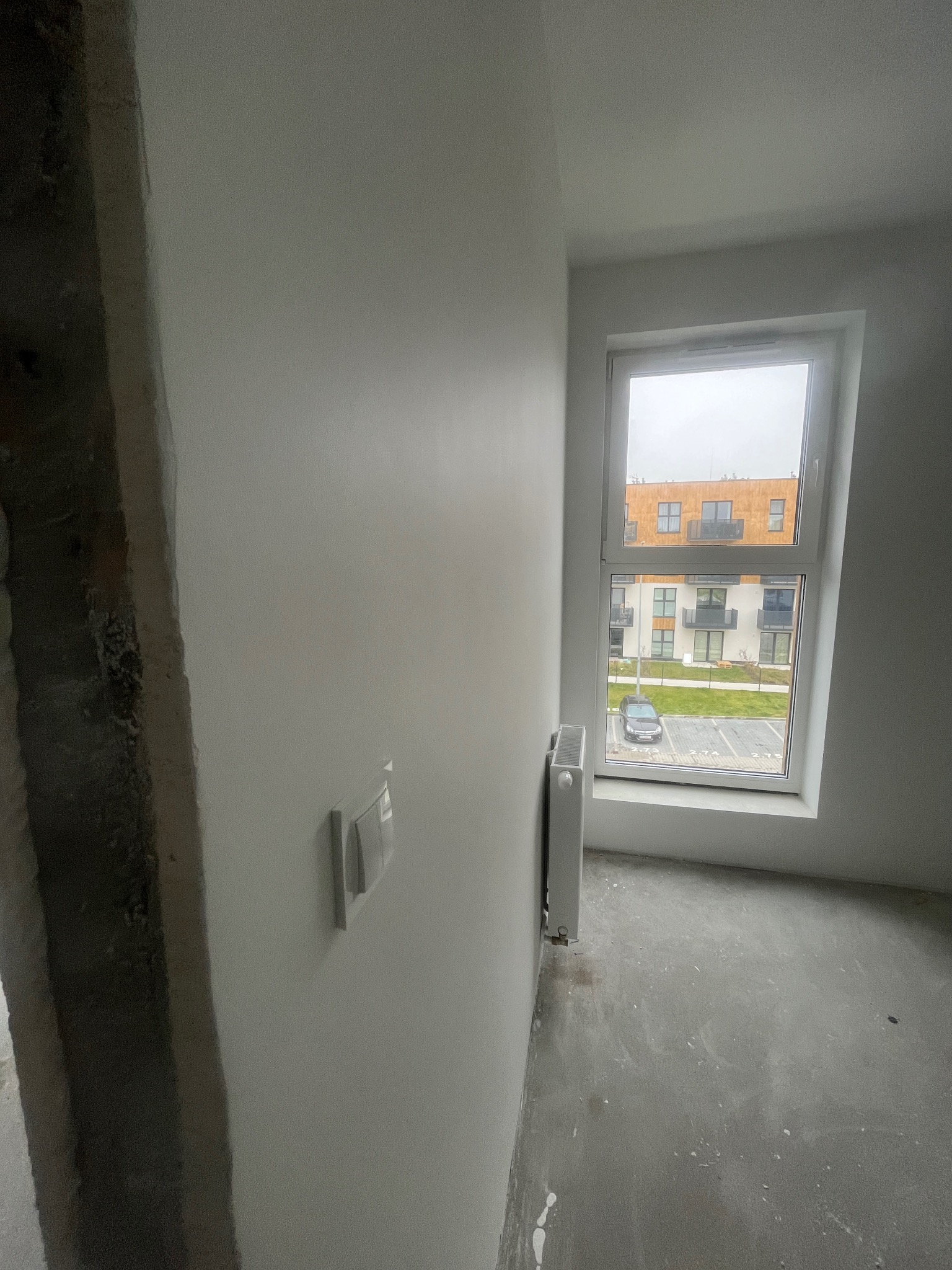 Wnętrze w trakcie remontu: świeżo pomalowane ściany na biało, widoczny grzejnik pod oknem z widokiem na parking i budynek mieszkalny. Ościeżnica surowa, bez wykończenia.