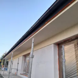 Świeżo otynkowana elewacja domu jednorodzinnego z czarną rynną, okna zabezpieczone folią, widoczne rusztowanie na tle błękitnego nieba.