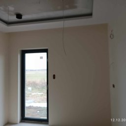 Sylwester Piotrowski - Pokój po szpachlowaniu, przygotowany do malowania, widoczne przewody wystające z sufitu i ślady po gniazdkach na ścianach, okno z widokiem na zewnątrz.