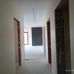 Sylwester Piotrowski - Korytarz w trakcie prac wykończeniowych, ściany po nałożeniu gładzi i wstępnym malowaniu, widoczne surowe ościeżnice drzwiowe i przygotowane podłoże pod posadzkę.