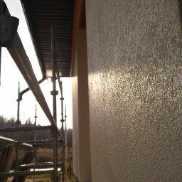 Sylwester Piotrowski - Świeżo nałożona, jasna struktura tynku na elewacji budynku w słoneczny dzień, widoczne rusztowanie i zabezpieczenie folią u podstawy ściany.