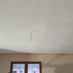 Sylwester Piotrowski - Wnętrze w trakcie prac w Piotrków Trybunalski: gładki, biały sufit po szpachlowaniu, z widocznymi wystającymi przewodami elektrycznymi i pomarańczową taśmą malarską zabezpieczającą krawędzie ścian.