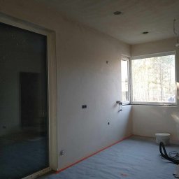 Sylwester Piotrowski - Pokój w trakcie remontu: ściany po szpachlowaniu, zabezpieczona podłoga, widoczne puszki elektryczne i okno z widokiem na drzewa.