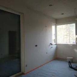 Pokój w trakcie remontu: ściany po szpachlowaniu, zabezpieczona podłoga, widoczne puszki elektryczne i okno z widokiem na drzewa.