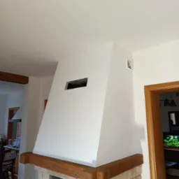 Biały kominek z drewnianą belką i kamiennym wykończeniem, widoczny w częściowo wykończonym wnętrzu z drzwiami po prawej stronie i fragmentem kuchni w tle.