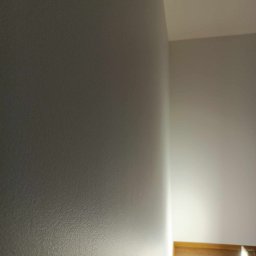 Sylwester Piotrowski - Szara ściana wewnętrzna z widoczną fakturą, oświetlona od dołu, z drewnianą podłogą w tle.