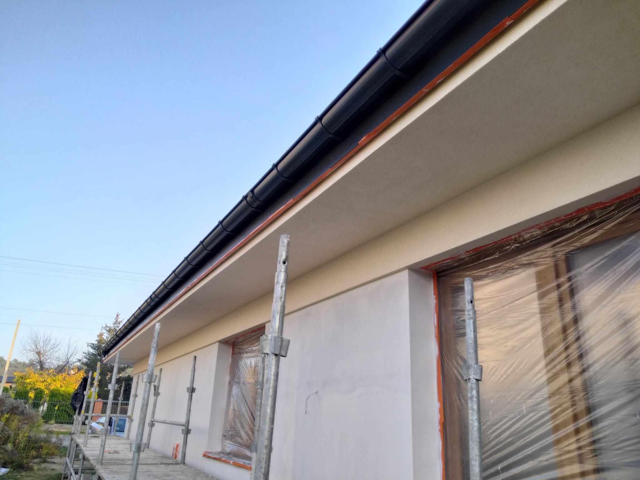 Świeżo otynkowana elewacja domu jednorodzinnego z czarną rynną, okna zabezpieczone folią, widoczne rusztowanie na tle błękitnego nieba.