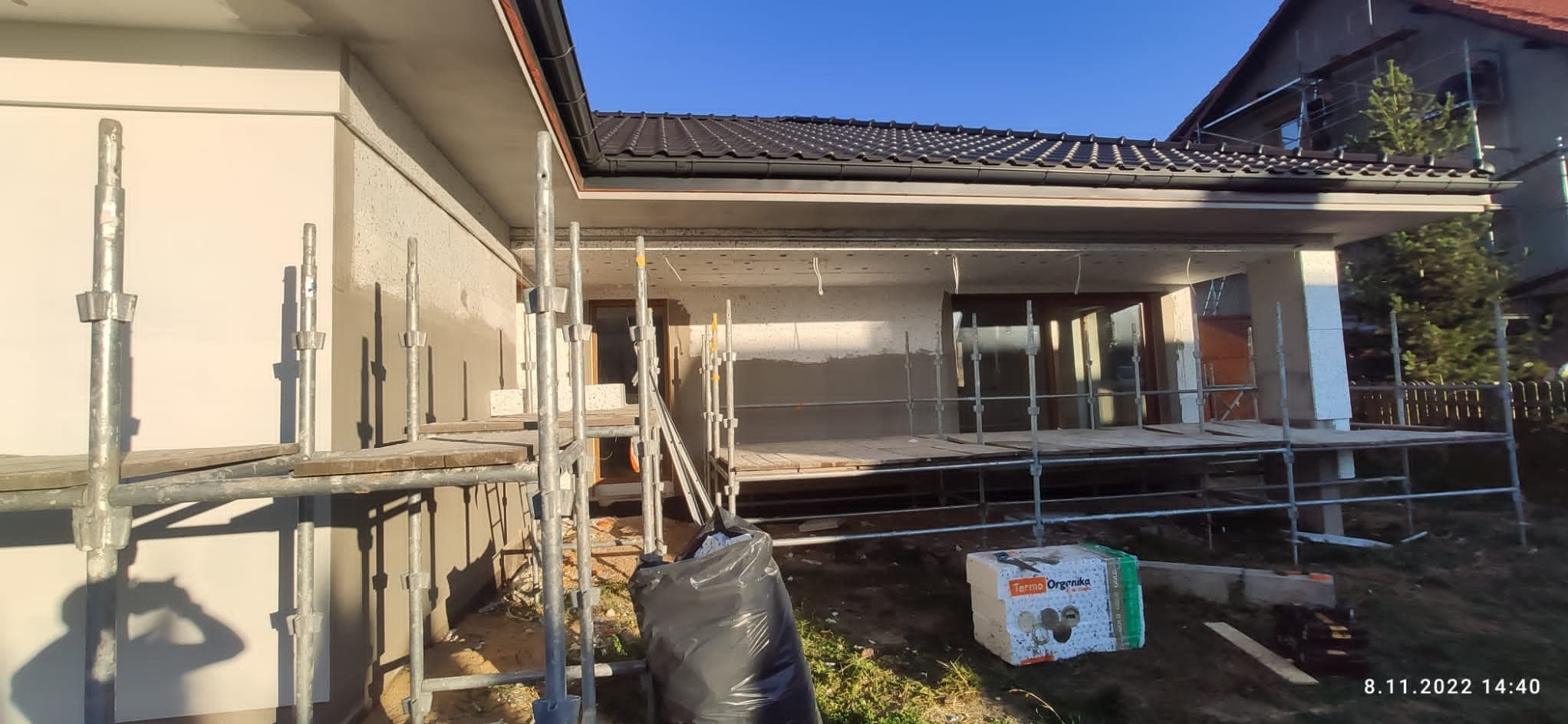 Dom w trakcie prac, z widocznymi rusztowaniami, ocieplonymi ścianami i częściowo wykonanymi tynkami zewnętrznymi, na trawniku leży paczka styropianu termo organika oraz worek z odpadami budowlanymi.