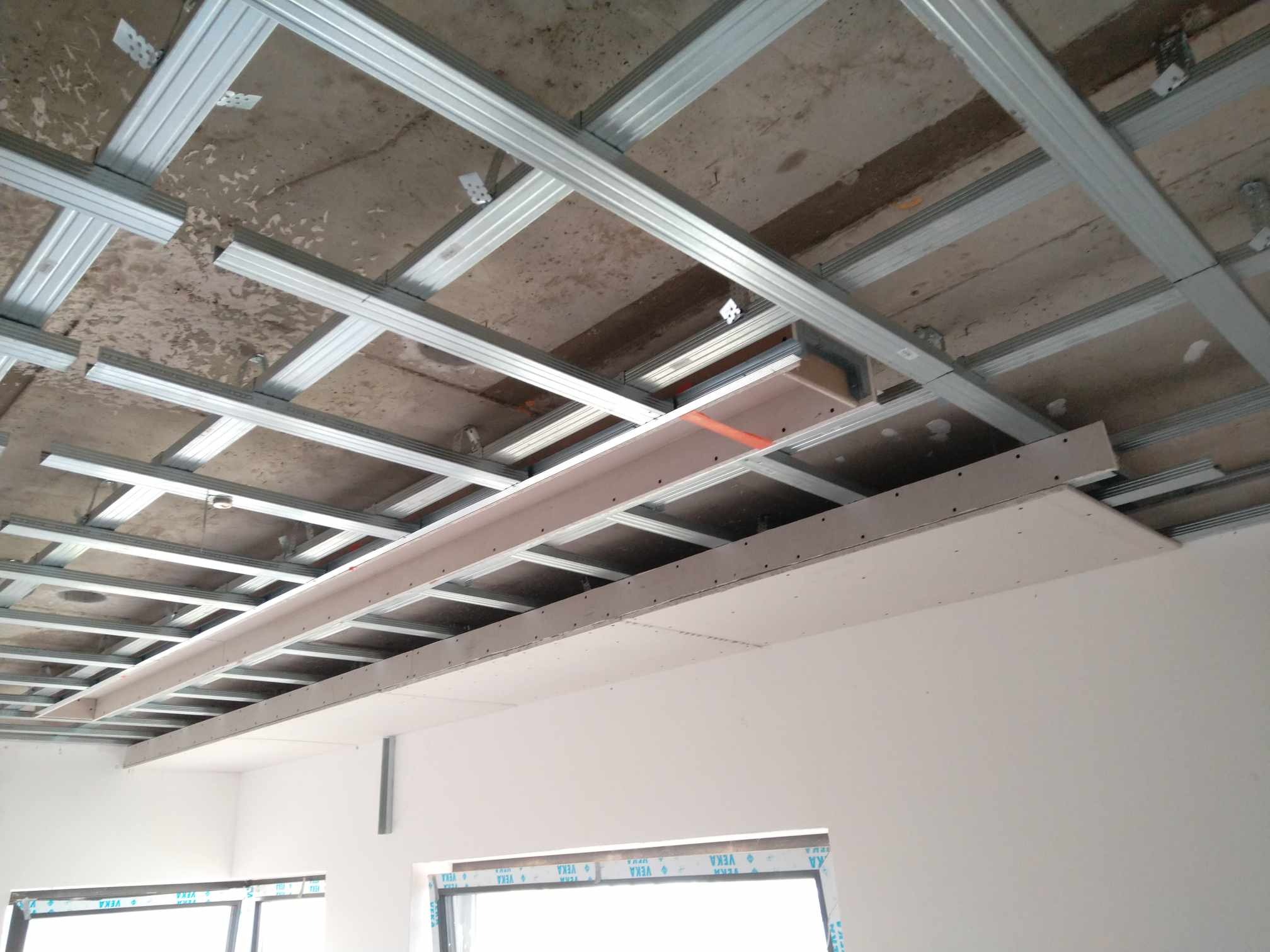 Montaż sufitu podwieszanego z płyty gipsowo-kartonowej, widoczne metalowe profile i częściowo zamontowana płyta, surowy betonowy strop w tle.