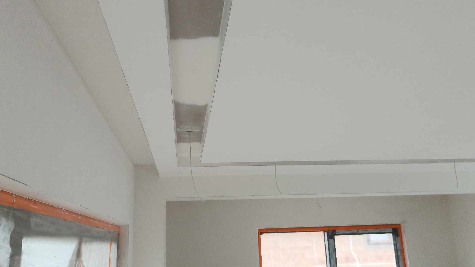 Nowoczesny sufit podwieszany z wnęką na oświetlenie, świeżo pomalowane ściany i okna zabezpieczone folią malarską.