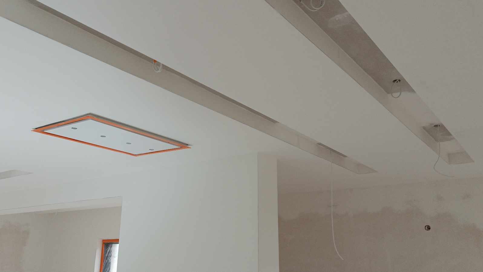 Sufit podwieszany z pomarańczową ramką i oświetleniem LED, widoczne listwy gipsowe na suficie, ściany w trakcie wykończenia.