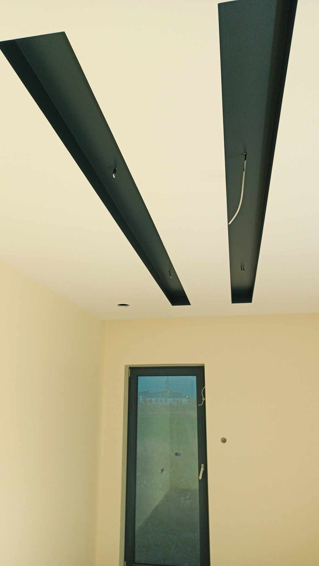 Dwa czarne, prostokątne elementy dekoracyjne zamontowane na białym suficie w nowym pomieszczeniu z beżowymi ścianami i czarnymi drzwiami balkonowymi.