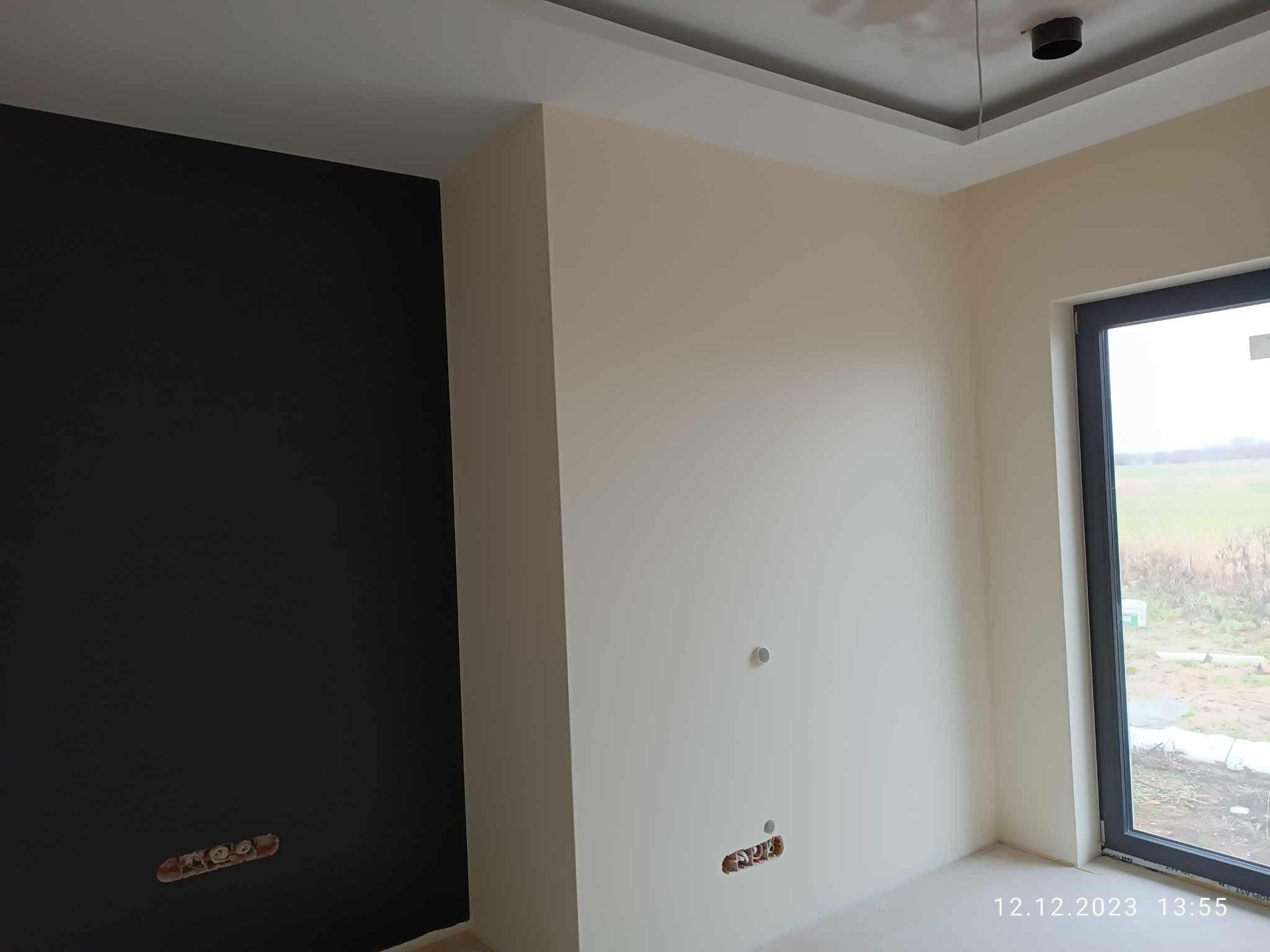 Świeżo pomalowane wnętrze z czarną ścianą kontrastującą z kremowymi ścianami, widoczne gniazdka elektryczne bez osprzętu i sufit z zamontowanym oświetleniem punktowym.