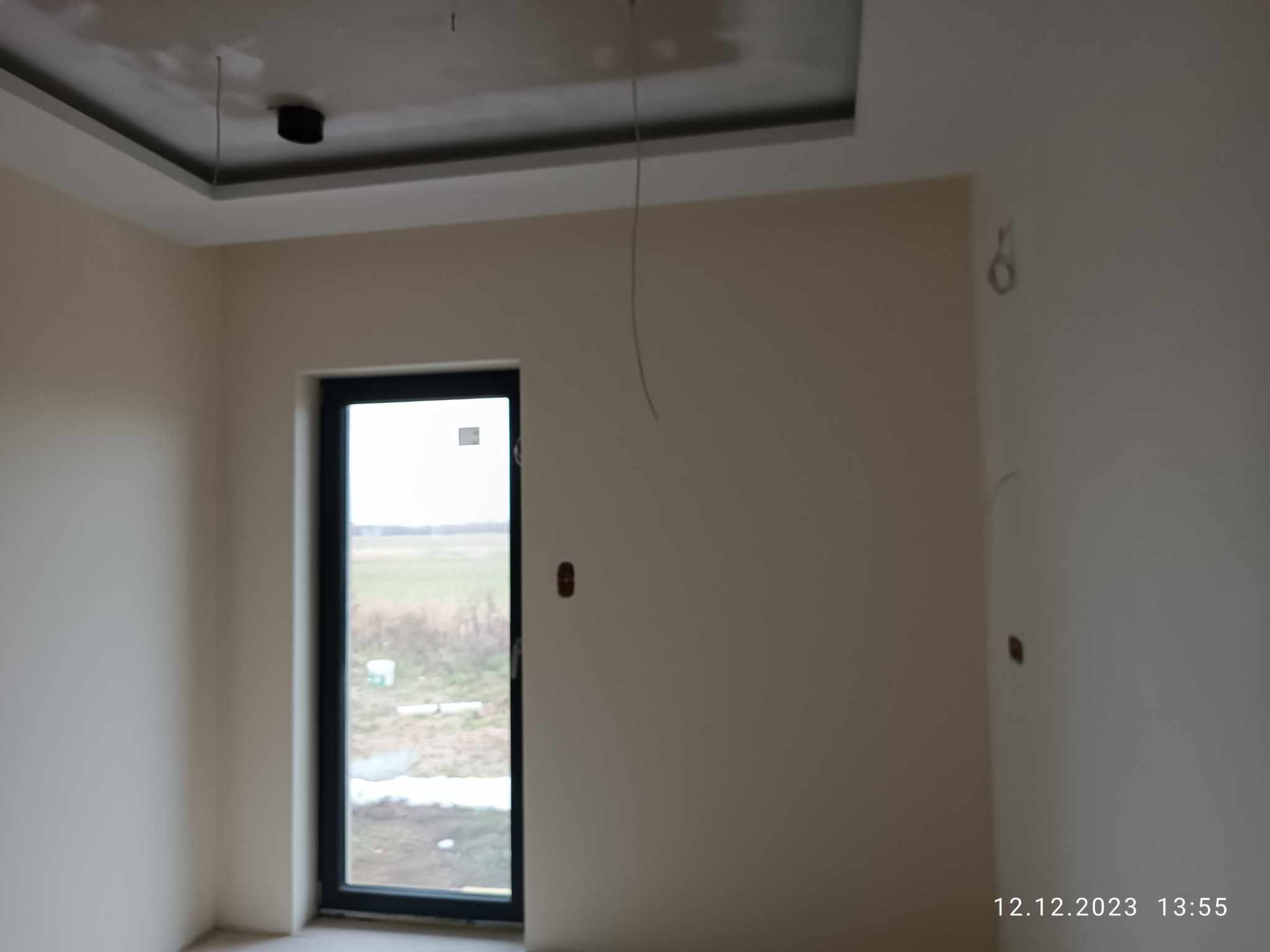 Pokój po szpachlowaniu, przygotowany do malowania, widoczne przewody wystające z sufitu i ślady po gniazdkach na ścianach, okno z widokiem na zewnątrz.