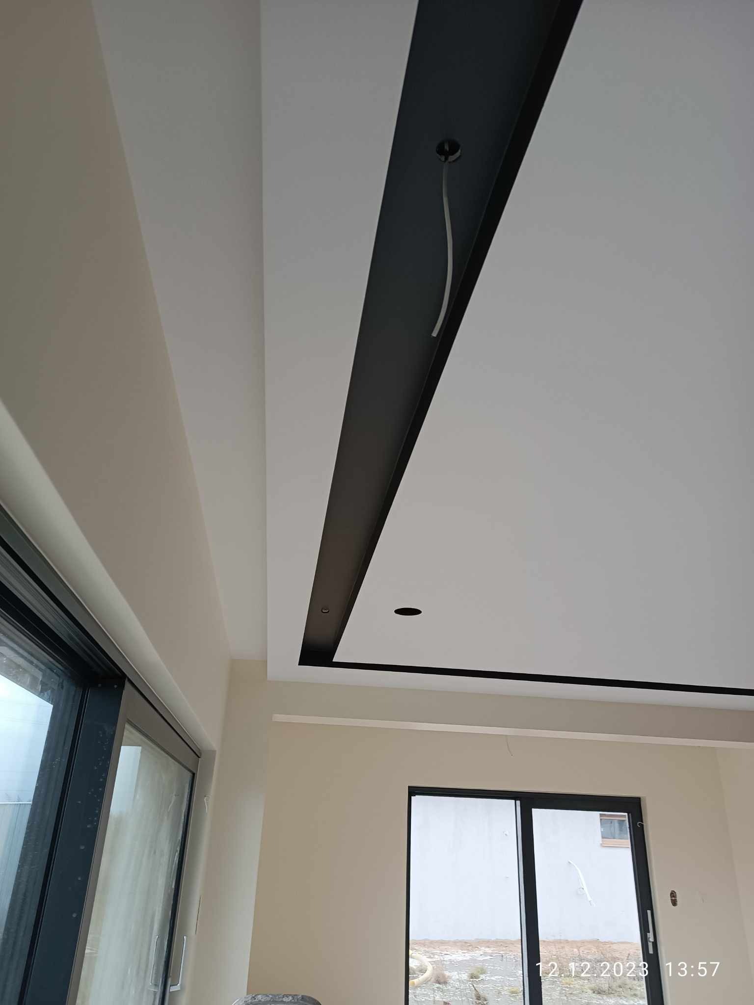 Nowoczesny sufit podwieszany z czarną listwą dekoracyjną i punktowym oświetleniem, widok z dołu na tle jasnych ścian i okna.