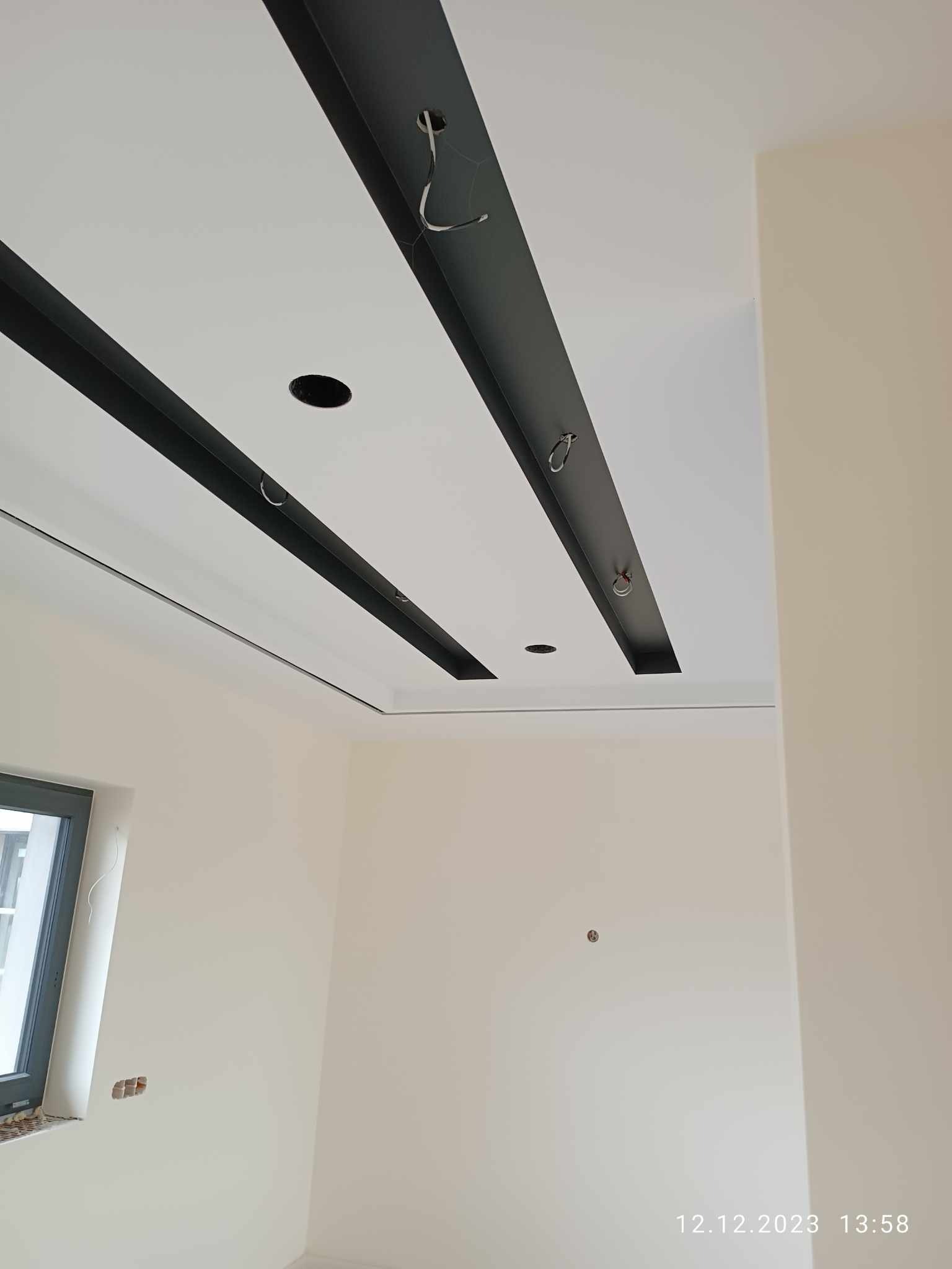 Nowoczesny sufit podwieszany z czarnymi elementami dekoracyjnymi i otworami na oświetlenie w jasnym wnętrzu w trakcie montażu.