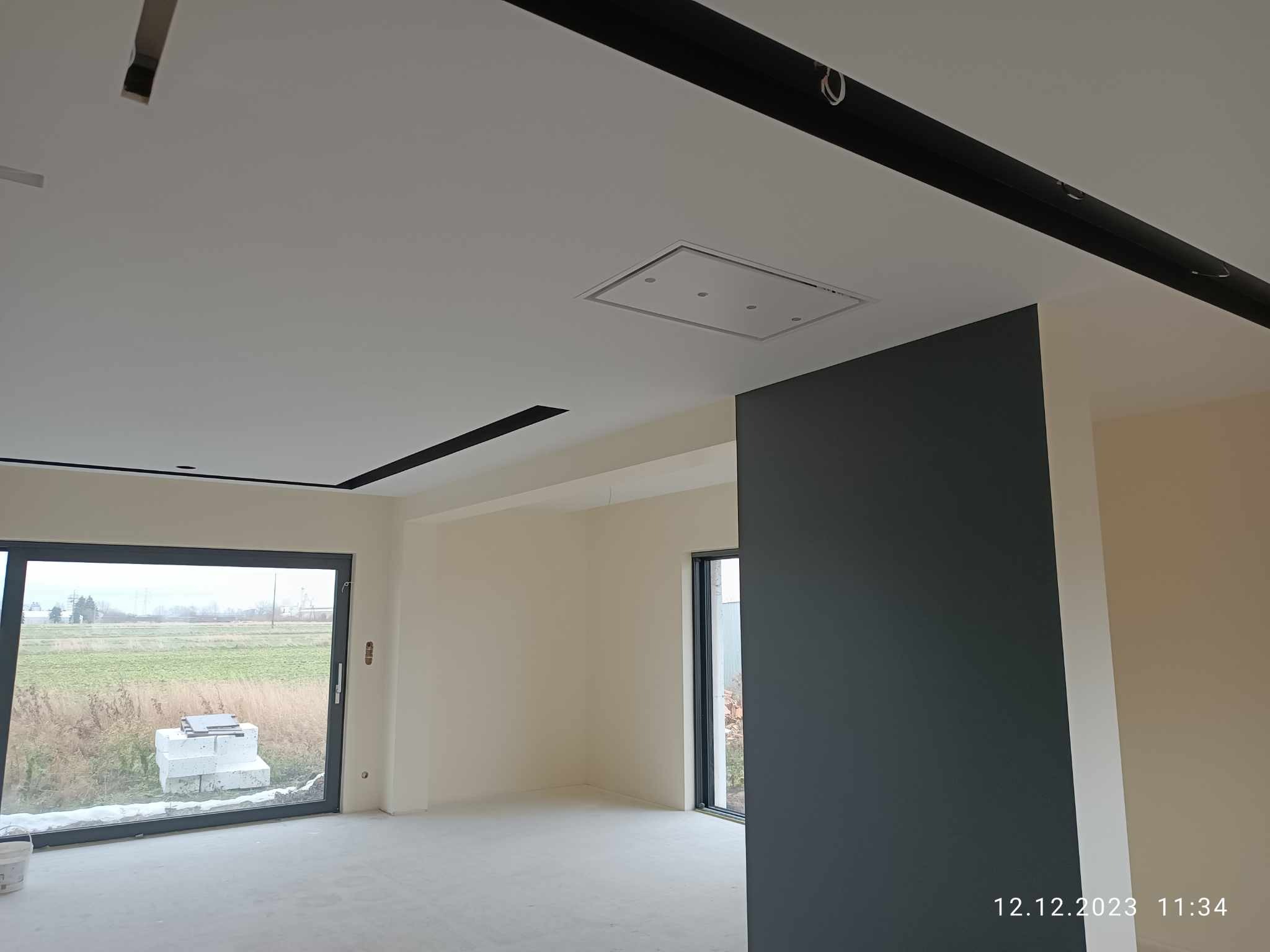 Wnętrze w trakcie wykańczania: gładkie, jasne ściany, sufit z wbudowanym panelem oświetleniowym, widoczne elementy konstrukcyjne w kolorze czarnym, duże okno z widokiem na zewnątrz.