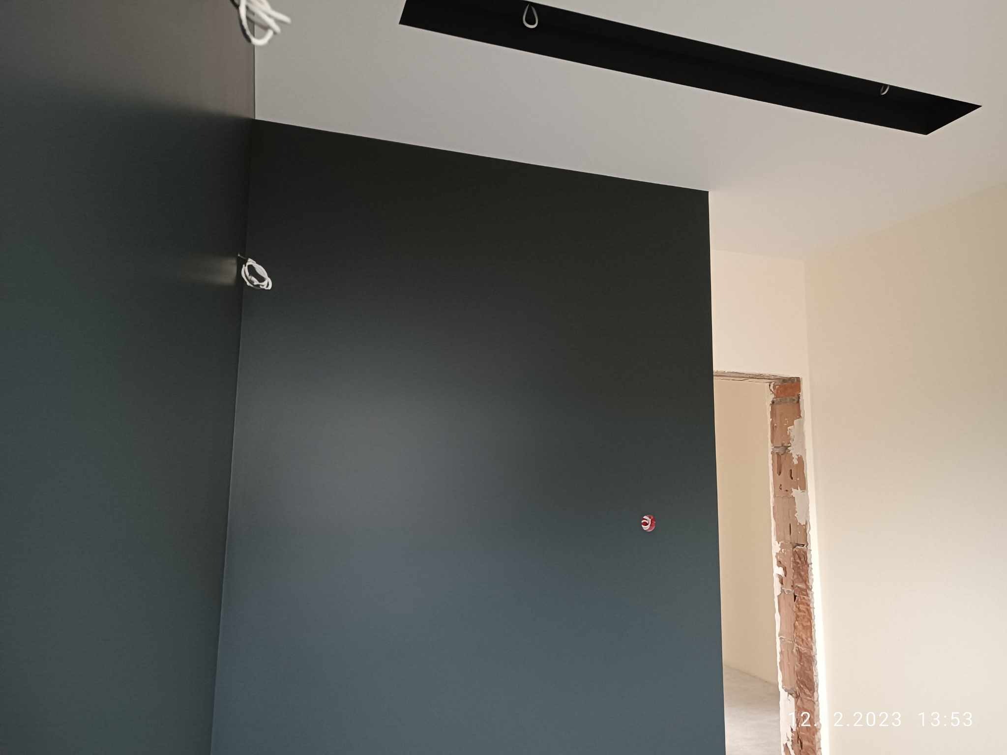 Świeżo pomalowana, ciemnoszara ściana w rogu pokoju z widoczną instalacją elektryczną i czarnym, prostokątnym elementem oświetleniowym na suficie. W tle widoczne surowe wykończenie otworu drzwiowego.