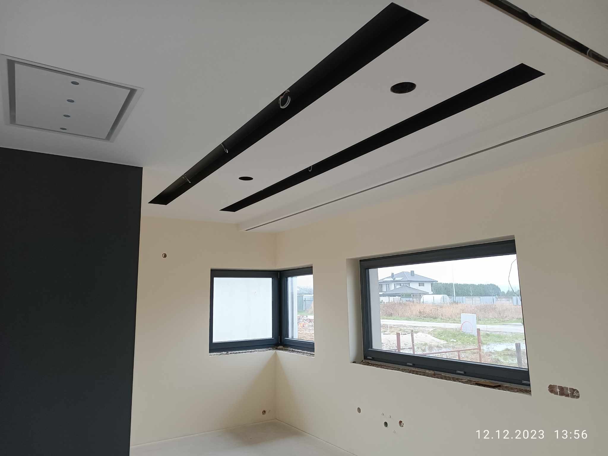 Wnętrze w trakcie prac wykończeniowych: biały sufit z czarnymi, prostokątnymi elementami dekoracyjnymi i wbudowanym oświetleniem, jasne ściany z dwoma szarymi oknami, jedna ściana pomalowana...