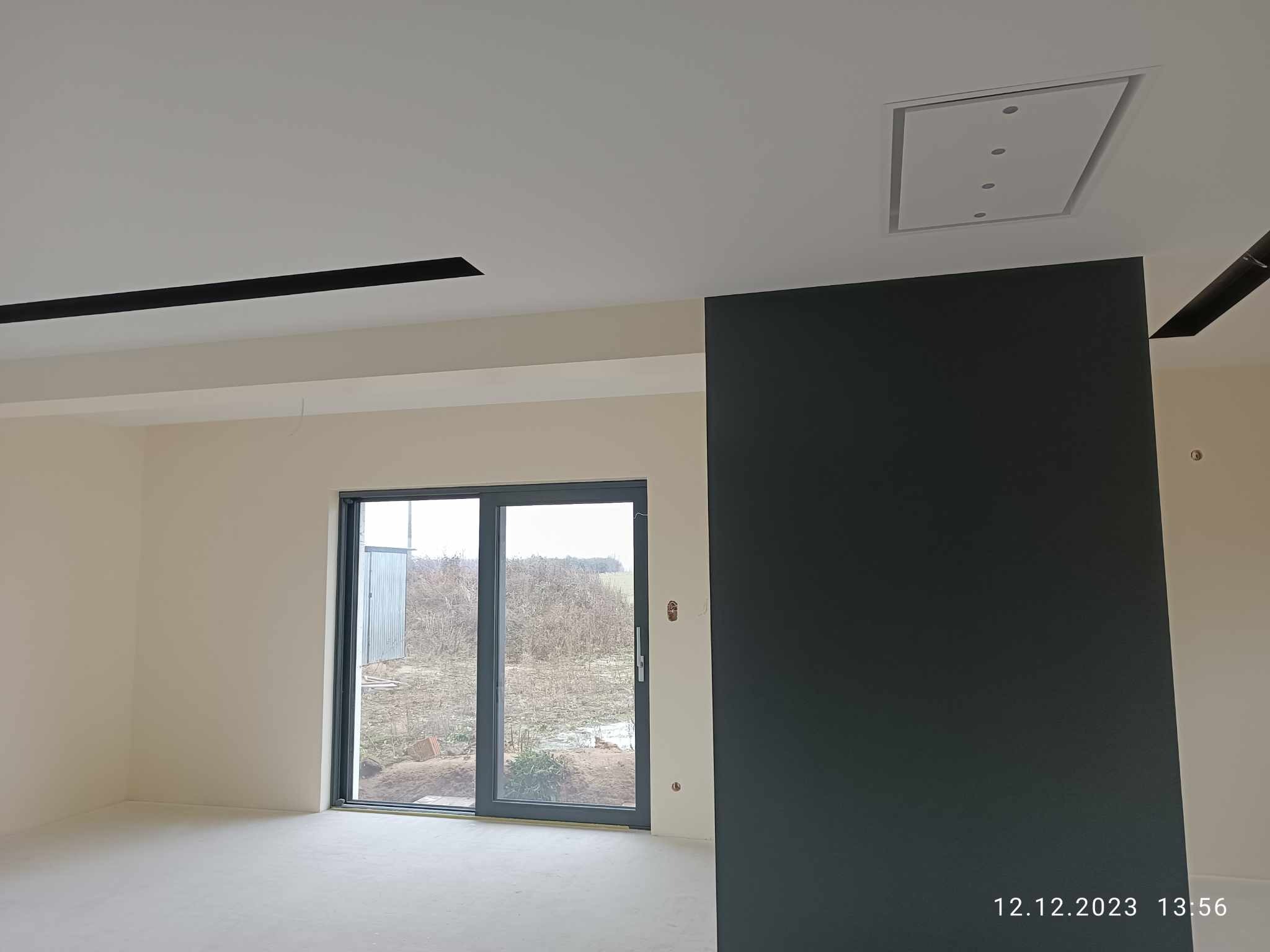 Wnętrze w stanie deweloperskim z dużym oknem balkonowym, ścianą pomalowaną na ciemny kolor i elementami dekoracyjnymi na suficie.