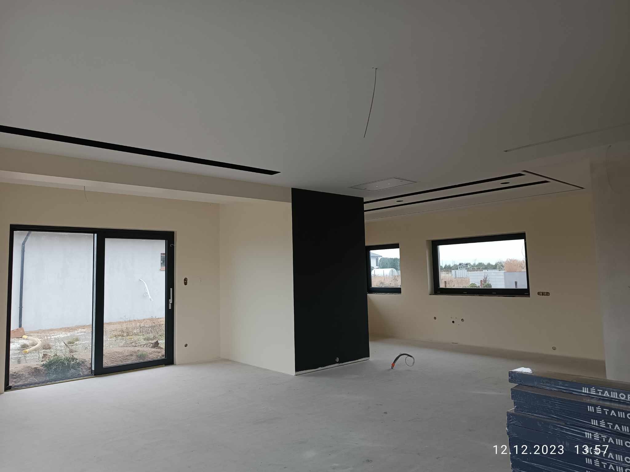 Wnętrze w trakcie prac wykończeniowych: surowa podłoga, ściany pomalowane na kremowo z czarnym panelem, czarne obramowania okien i sufitu, widoczne okna i drzwi balkonowe.