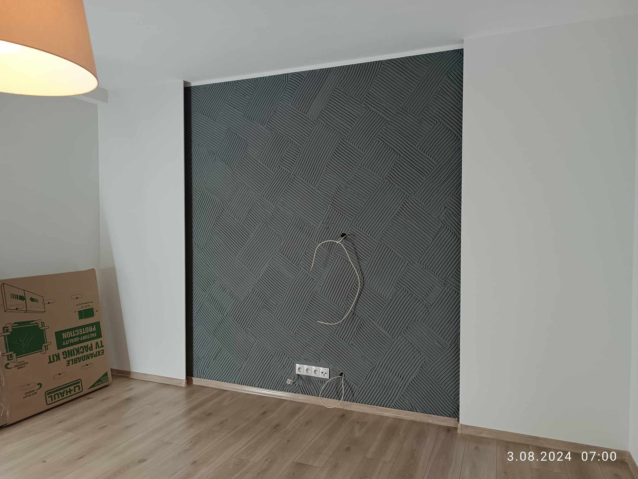 Ściana z dekoracyjną szarą strukturą przypominającą ułożone cegły, z widocznymi gniazdkami elektrycznymi i wystającymi kablami, świeżo położona podłoga, kartonowe pudło w rogu pomieszczenia