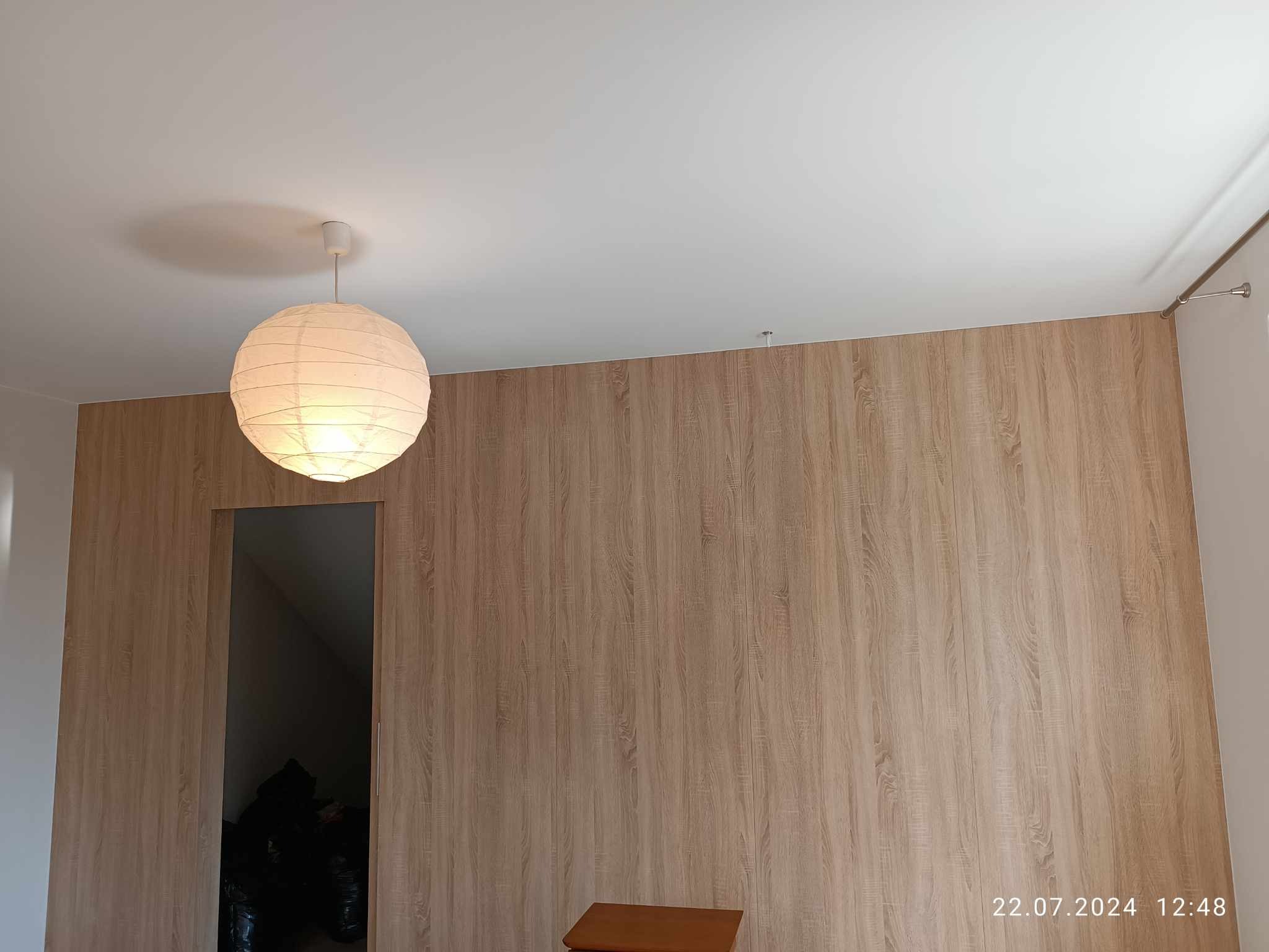 Pokój z wykończoną ścianą imitującą drewno, oświetlony okrągłą lampą papierową, z widocznym wejściem do ciemnego pomieszczenia.