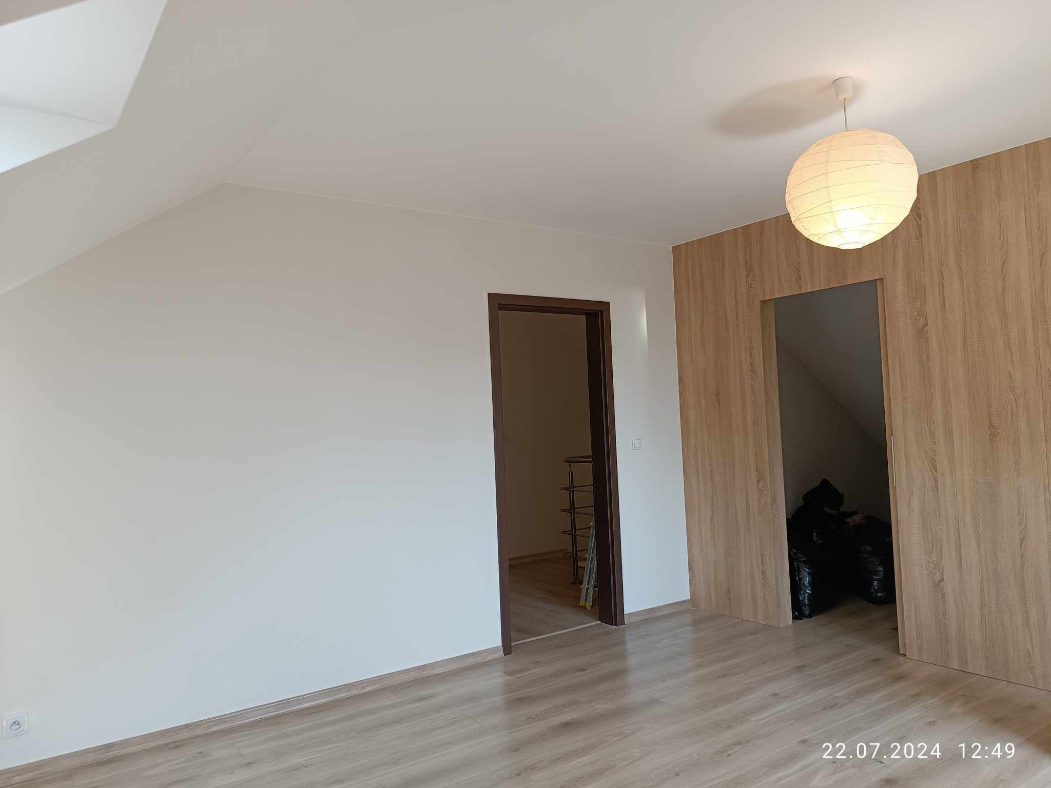 Poddasze po remoncie, ściany pomalowane na biało, podłoga wyłożona panelami, fragment ściany wykończony panelami imitującymi drewno, widoczne drzwi do kolejnego pomieszczenia oraz lampa wisząca.