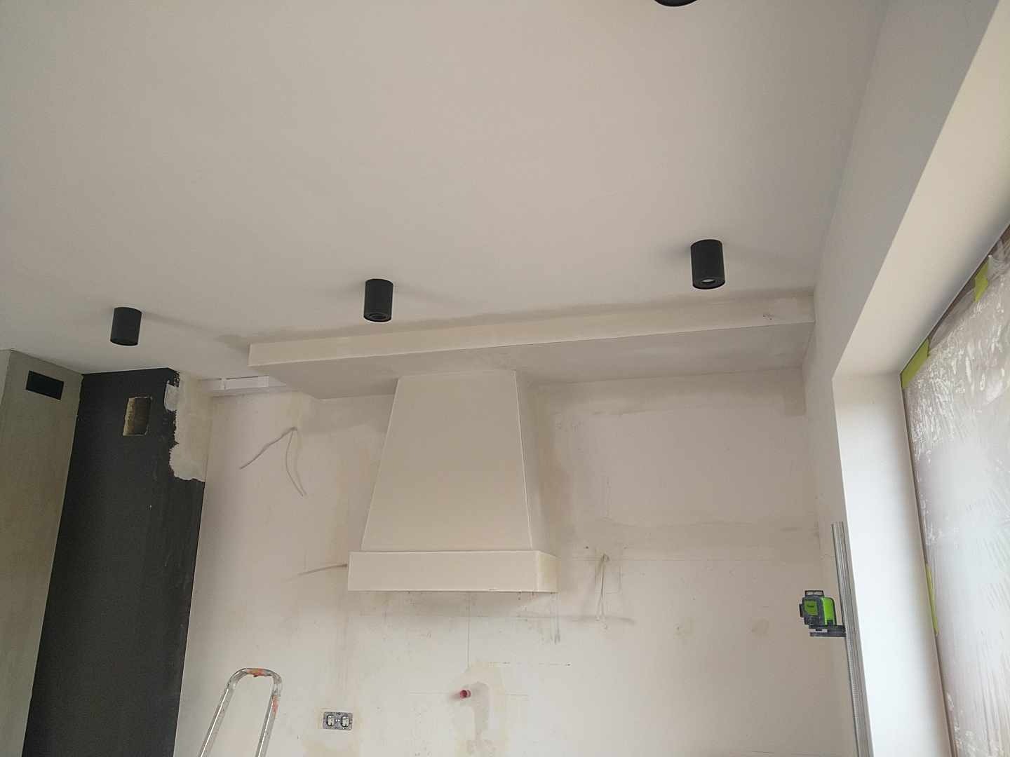 Wnętrze kuchni w trakcie prac wykończeniowych: ściany po szpachlowaniu, przygotowane do malowania, widoczne czarne lampy sufitowe, biały okap i poziomica laserowa.