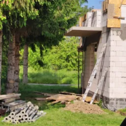 Wznoszony budynek z białych bloczków, widoczne szalunki, drabina oparta o ścianę, stosy podpór budowlanych na trawie, w tle zieleń drzew.