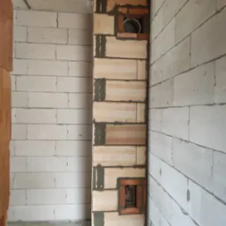 Wnętrze w trakcie budowy: fragment ściany z pustaków ceramicznych z otworem wentylacyjnym i kominowym, widoczne ślady zaprawy, ściany z białych bloczków.