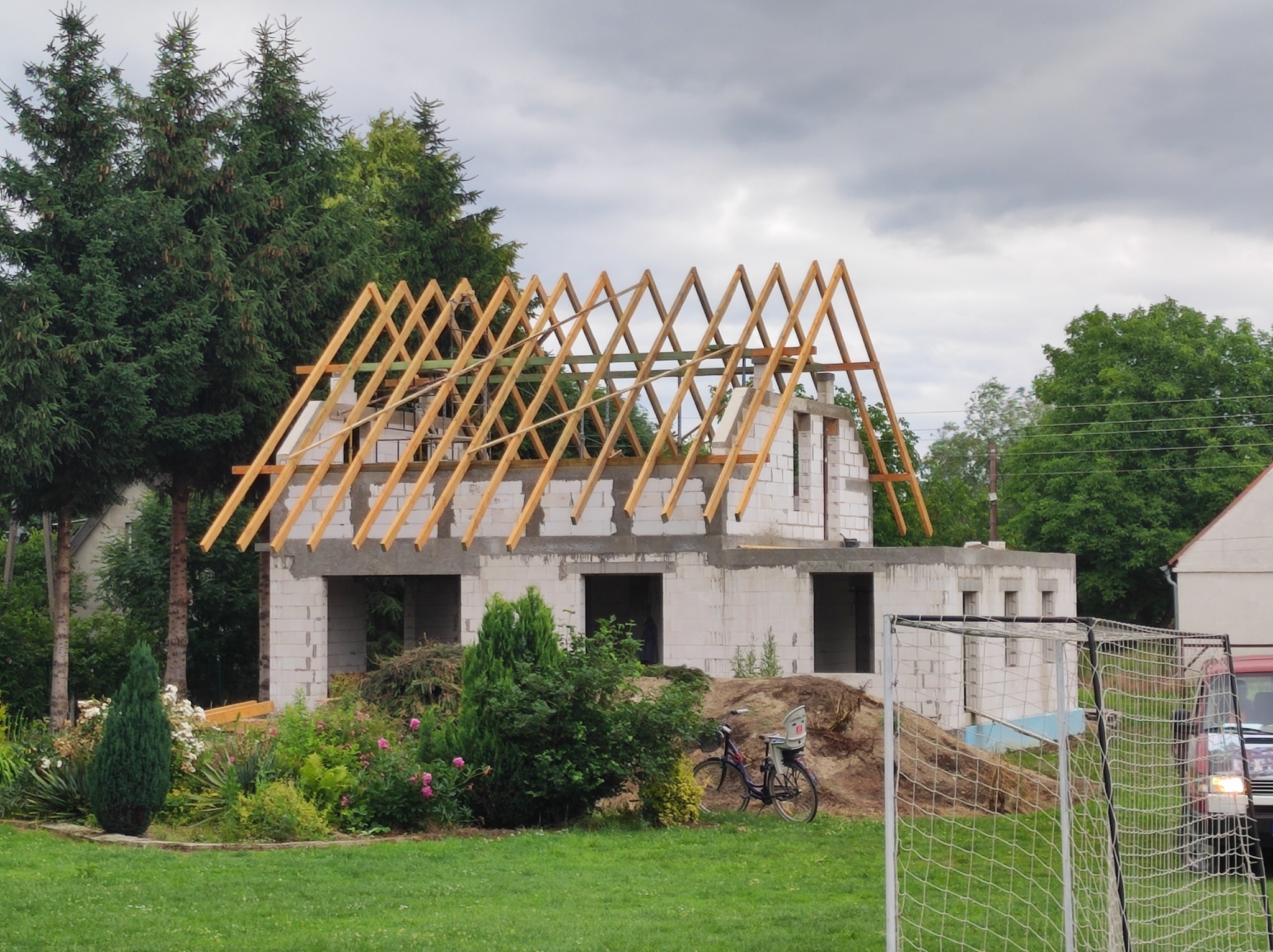 Surowy stan dwukondygnacyjnego budynku z białych pustaków, z drewnianą konstrukcją dachu w trakcie budowy, na tle zielonego ogrodu i zachmurzonego nieba.