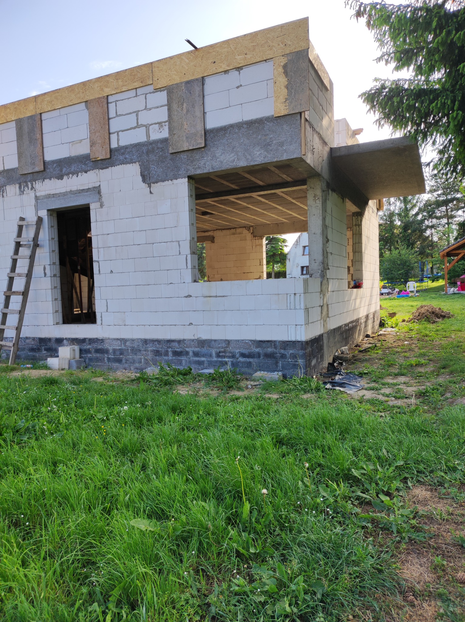 Surowy stan budynku z białych bloczków z widoczną konstrukcją stropu, drewnianymi deskami na górze i drabiną opartą o ścianę, otoczony zieloną trawą.