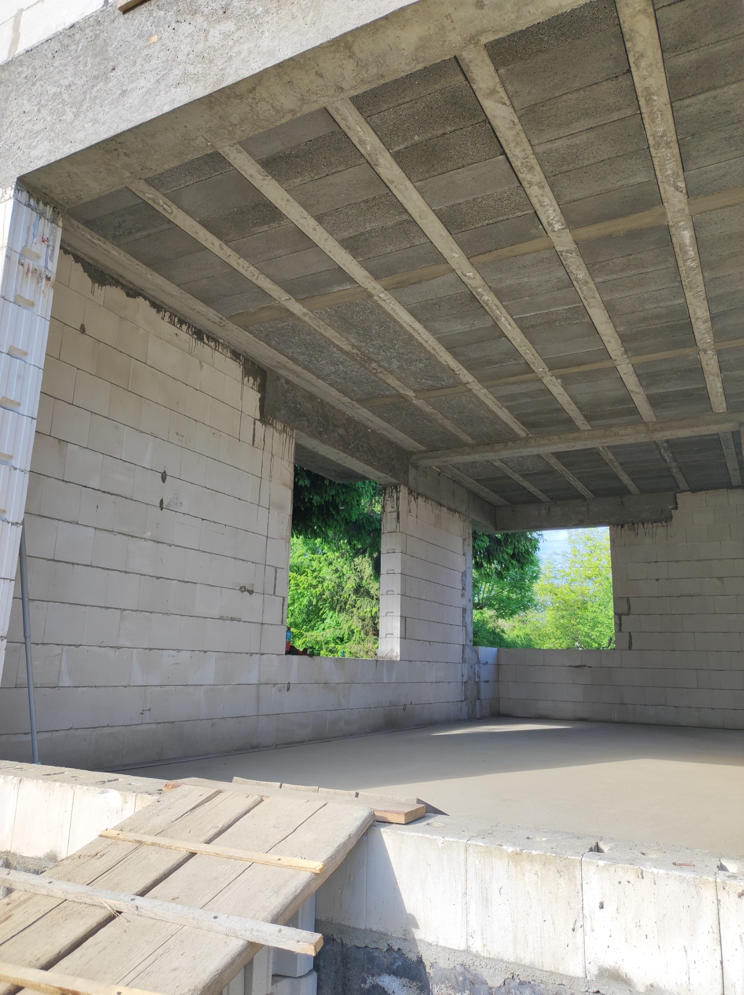 Wnętrze surowego domu w trakcie budowy, ściany z białych bloczków, betonowy strop z widocznymi belkami, widok przez otwory okienne na zieleń, drewniany podest na pierwszym planie.