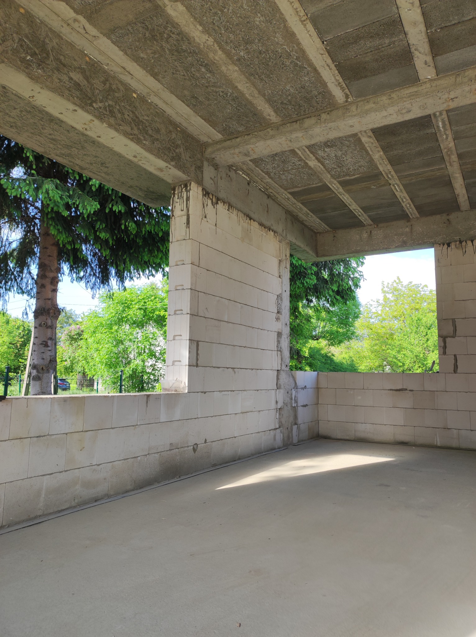 Surowy stan domu w trakcie budowy, ściany z betonu komórkowego, widoczne zbrojenia stropu, zieleń drzew w tle