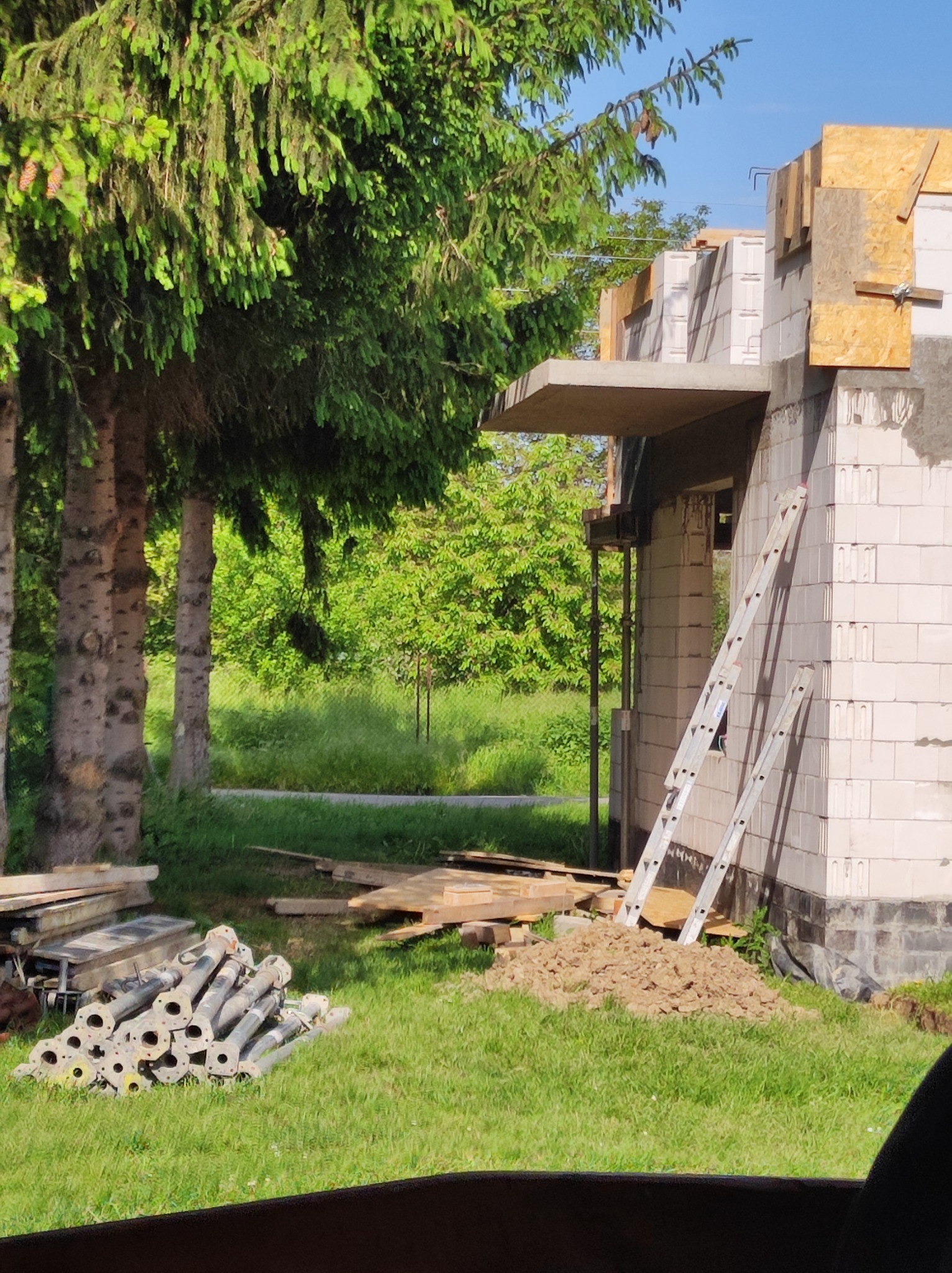 Wznoszony budynek z białych bloczków, widoczne szalunki, drabina oparta o ścianę, stosy podpór budowlanych na trawie, w tle zieleń drzew.