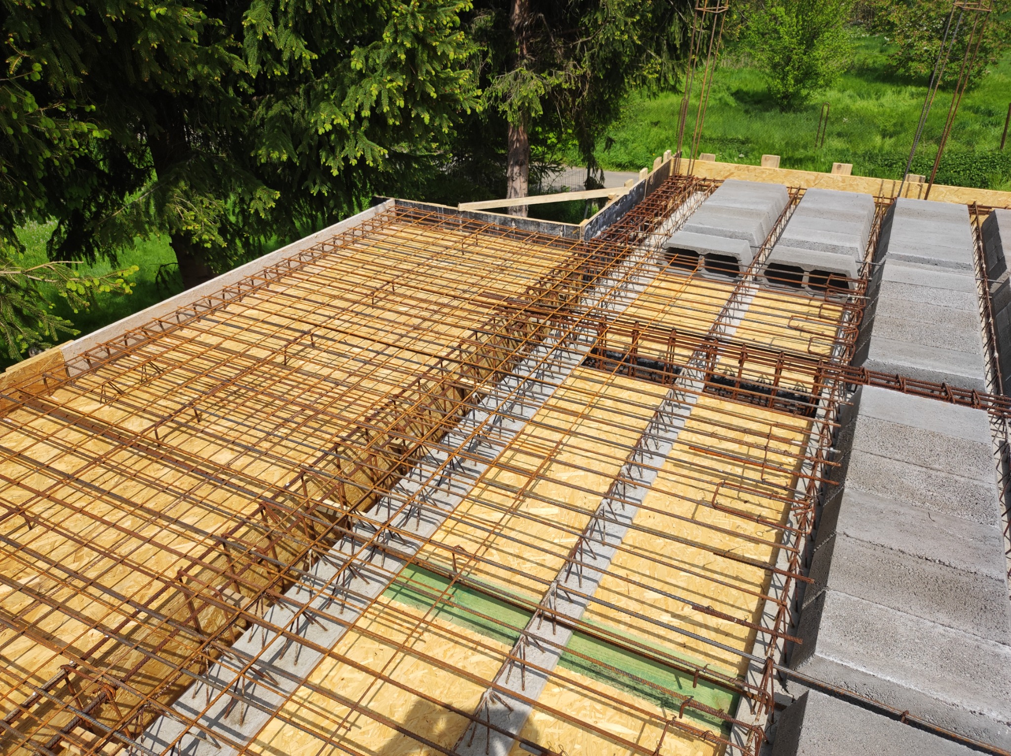 Wieniec stropowy w trakcie wykonywania: szalunek z płyty OSB, zbrojenie z prętów stalowych i pustaki szalunkowe, widok z góry na tle zieleni.