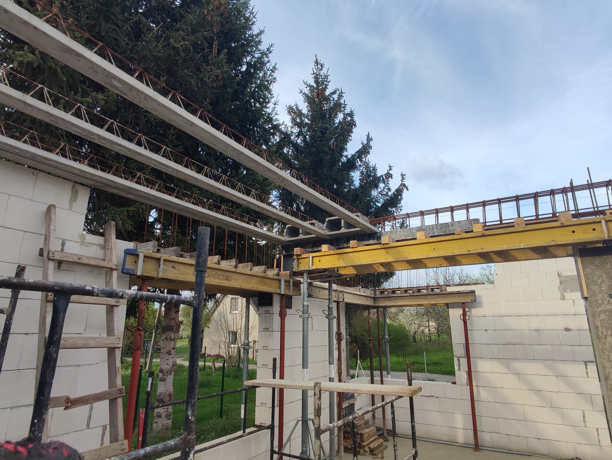 Wznoszenie ścian z bloczków, widoczne szalunki stropowe i zbrojenia, w tle drzewa i błękitne niebo.