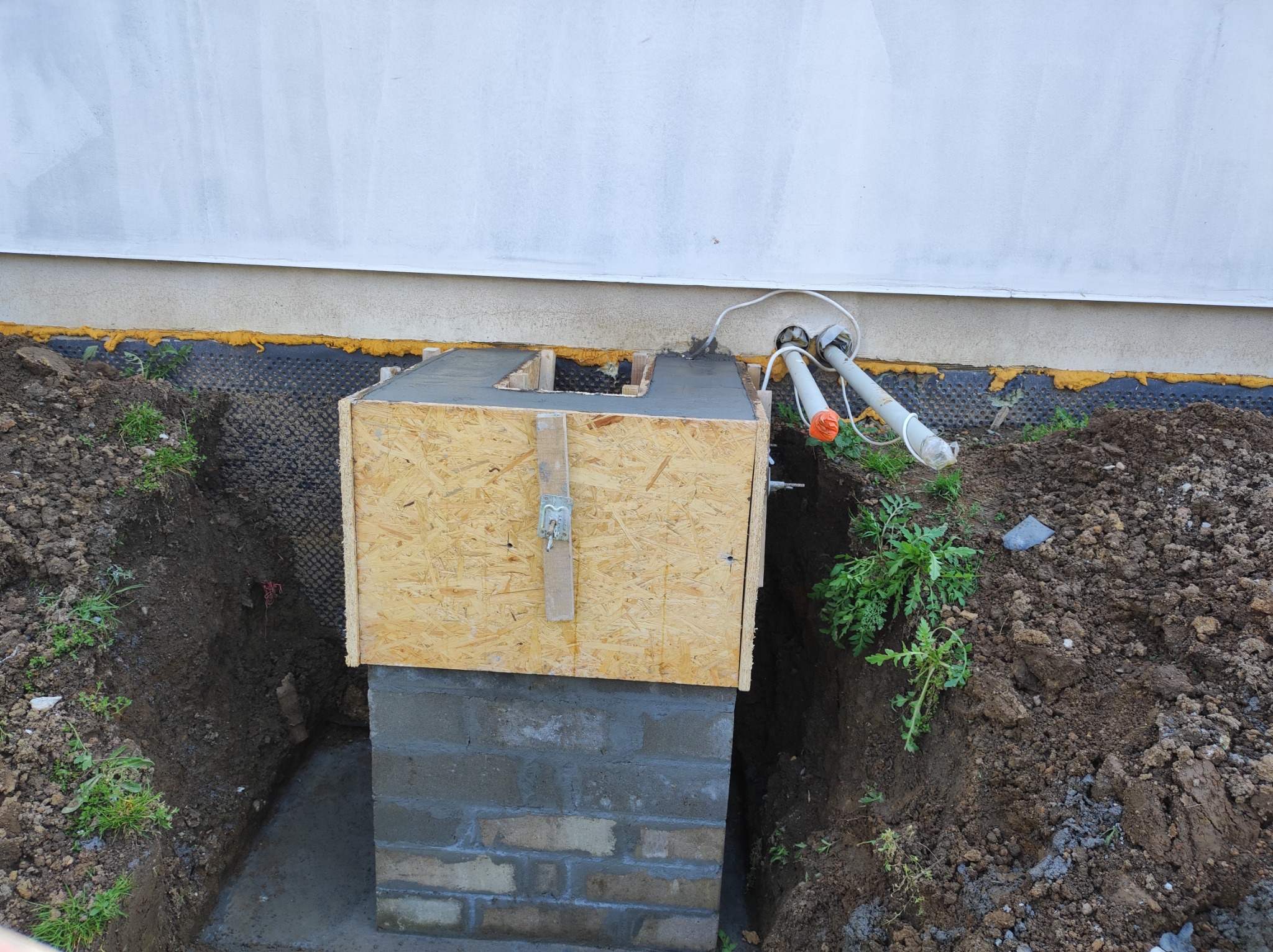 Wykop fundamentowy z murowanym fragmentem i drewnianym szalunkiem wypełnionym betonem, widoczne wystające rury i izolacja ściany budynku.