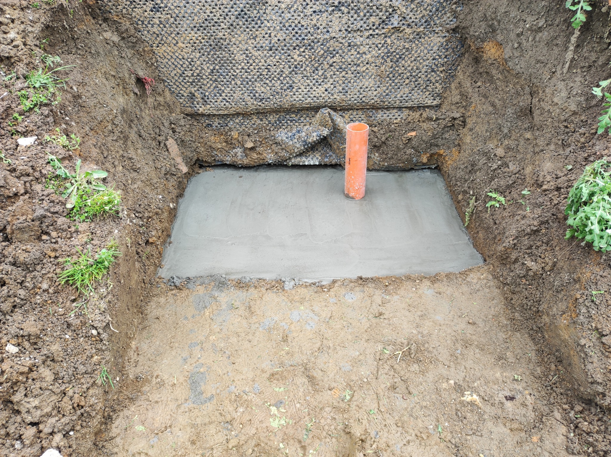 Wybetonowany fundament z wystającą rurą kanalizacyjną, zabezpieczony od strony gruntu czarną folią drenażową.