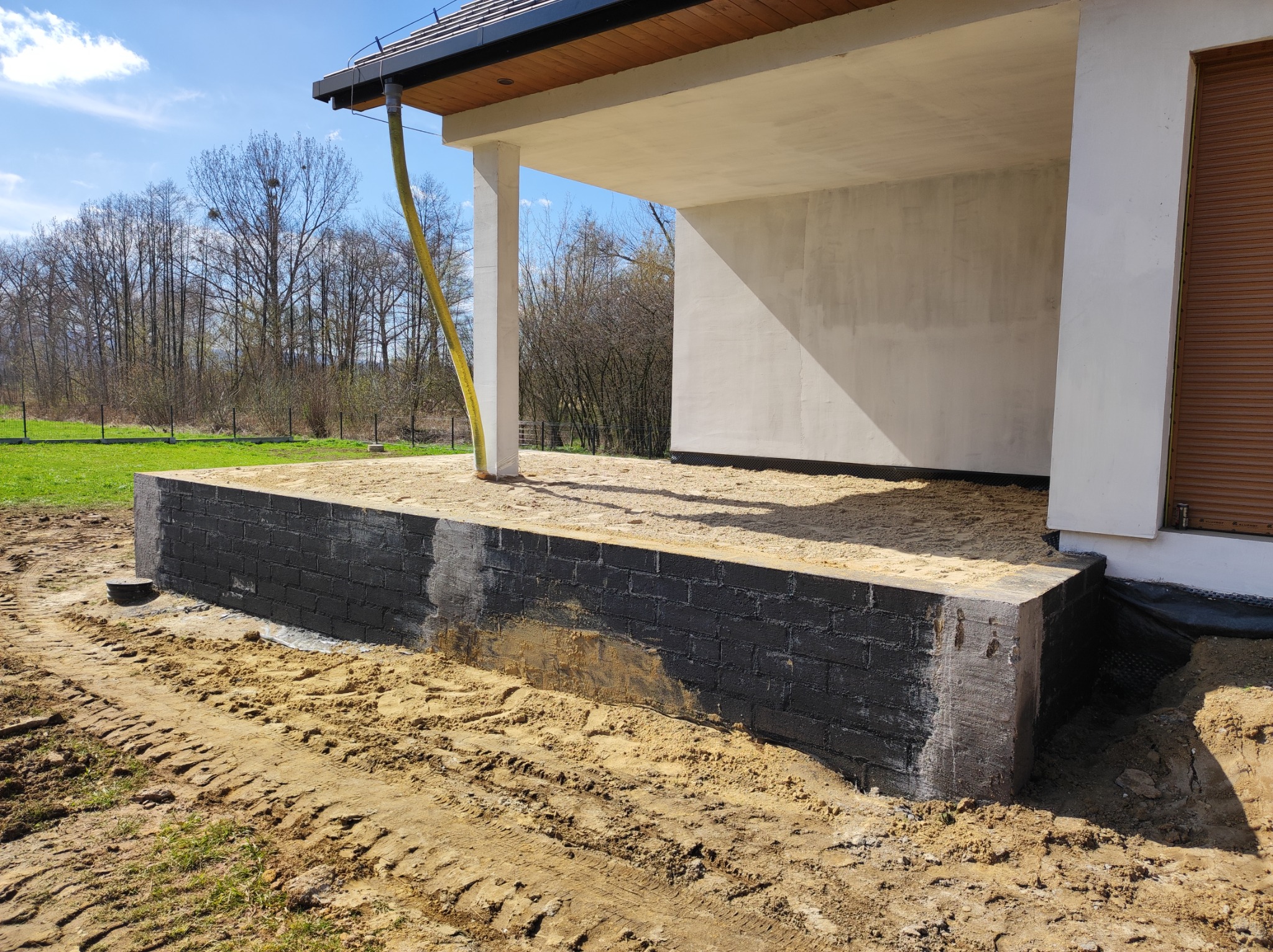 Fundamenty nowego domu z czarnych bloczków, częściowo zasypane piaskiem, widoczna żółta rura spustowa i fragment elewacji w surowym stanie.