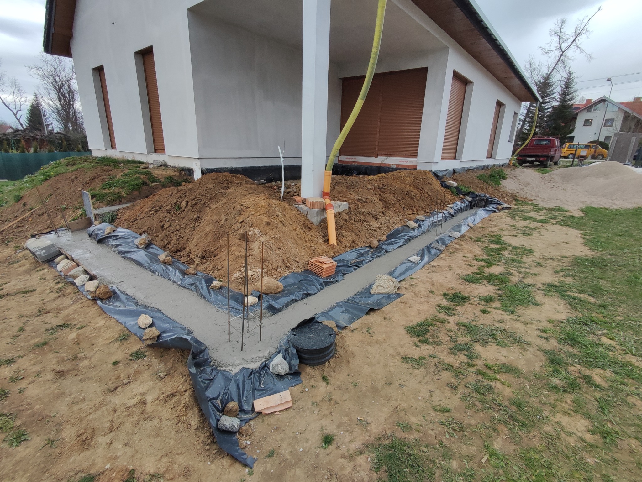 Wykop fundamentowy z zalanym betonem, zabezpieczony folią i kamieniami, widoczne pręty zbrojeniowe, rury kanalizacyjne i fragment budynku w tle.