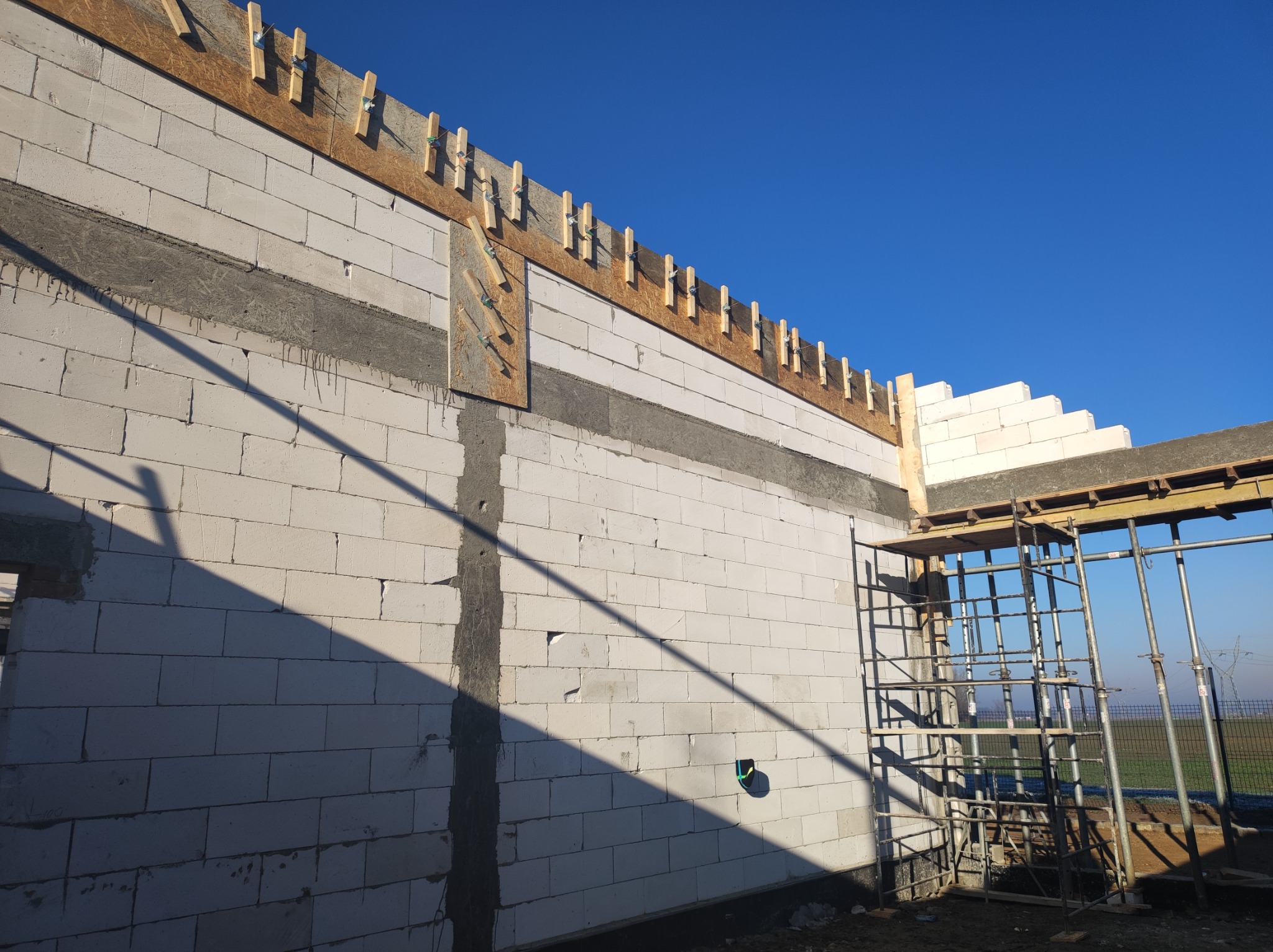 Wznoszenie ściany z białych bloczków z widocznym zbrojeniem poziomym i elementami szalunku stropu, na tle błękitnego nieba.