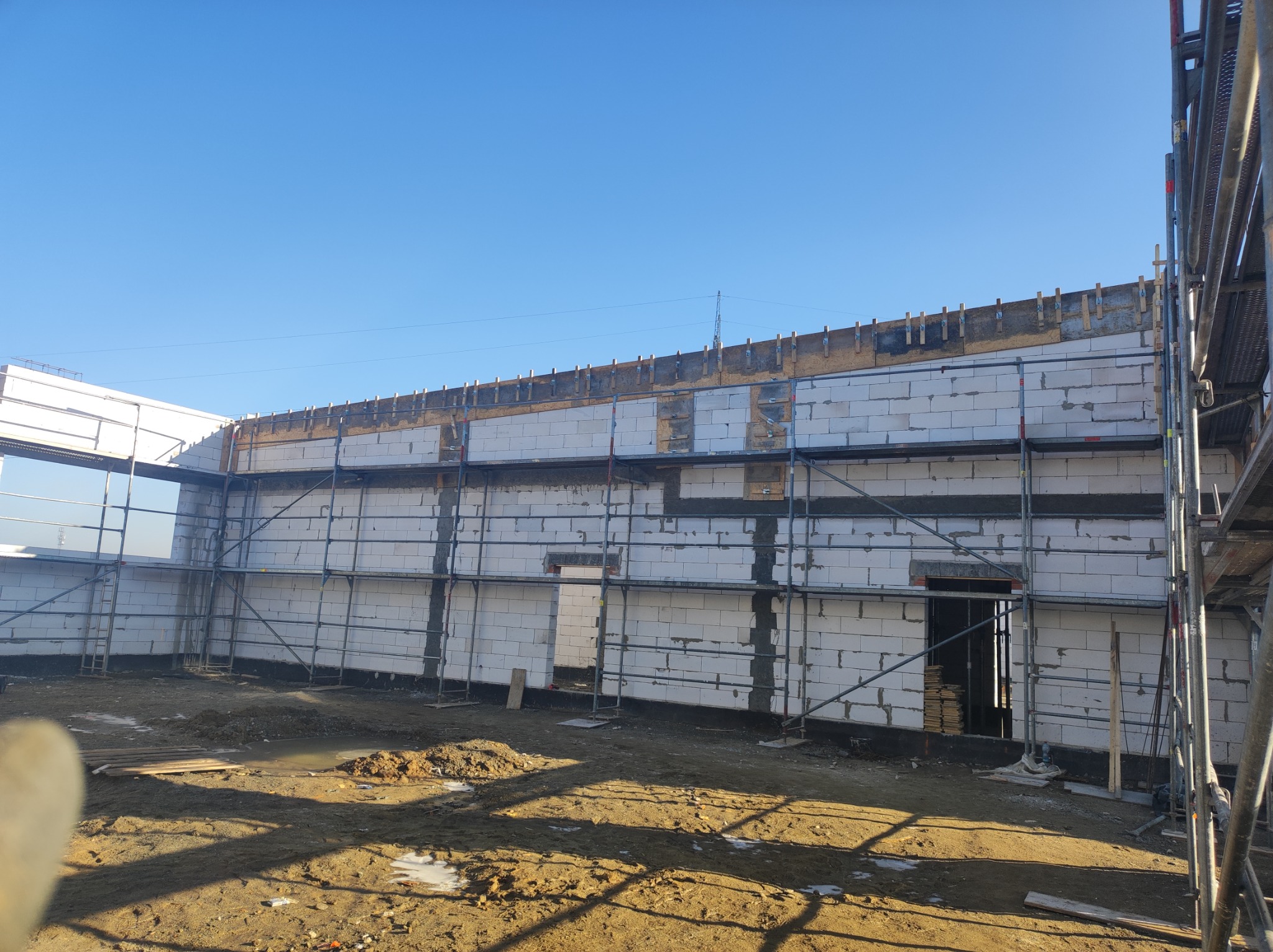 Wznoszenie ścian domu murowanego z białych bloczków, widoczne rusztowanie i szalunek stropowy, teren budowy w słoneczny dzień.