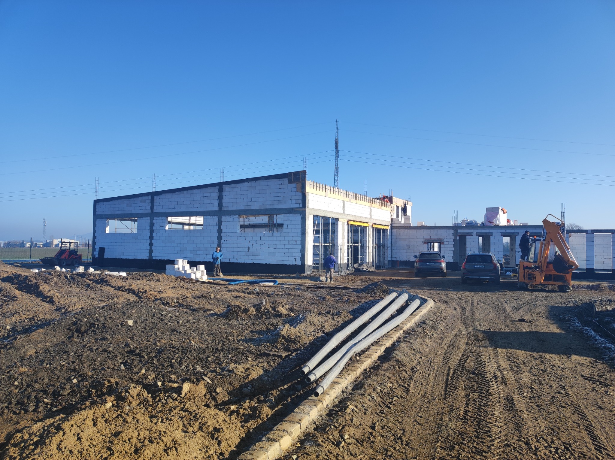 Wznoszenie ścian budynku z bloczków, widoczne rury w wykopie, maszyny budowlane i auta w tle, słoneczny dzień.