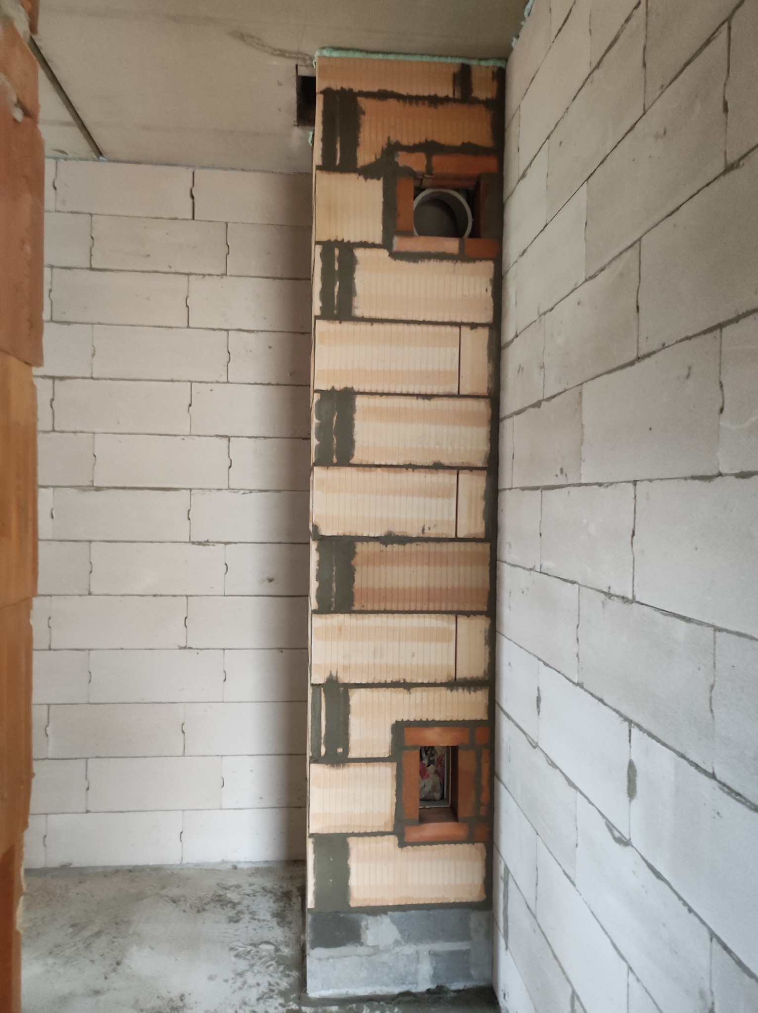 Wnętrze w trakcie budowy: fragment ściany z pustaków ceramicznych z otworem wentylacyjnym i kominowym, widoczne ślady zaprawy, ściany z białych bloczków.