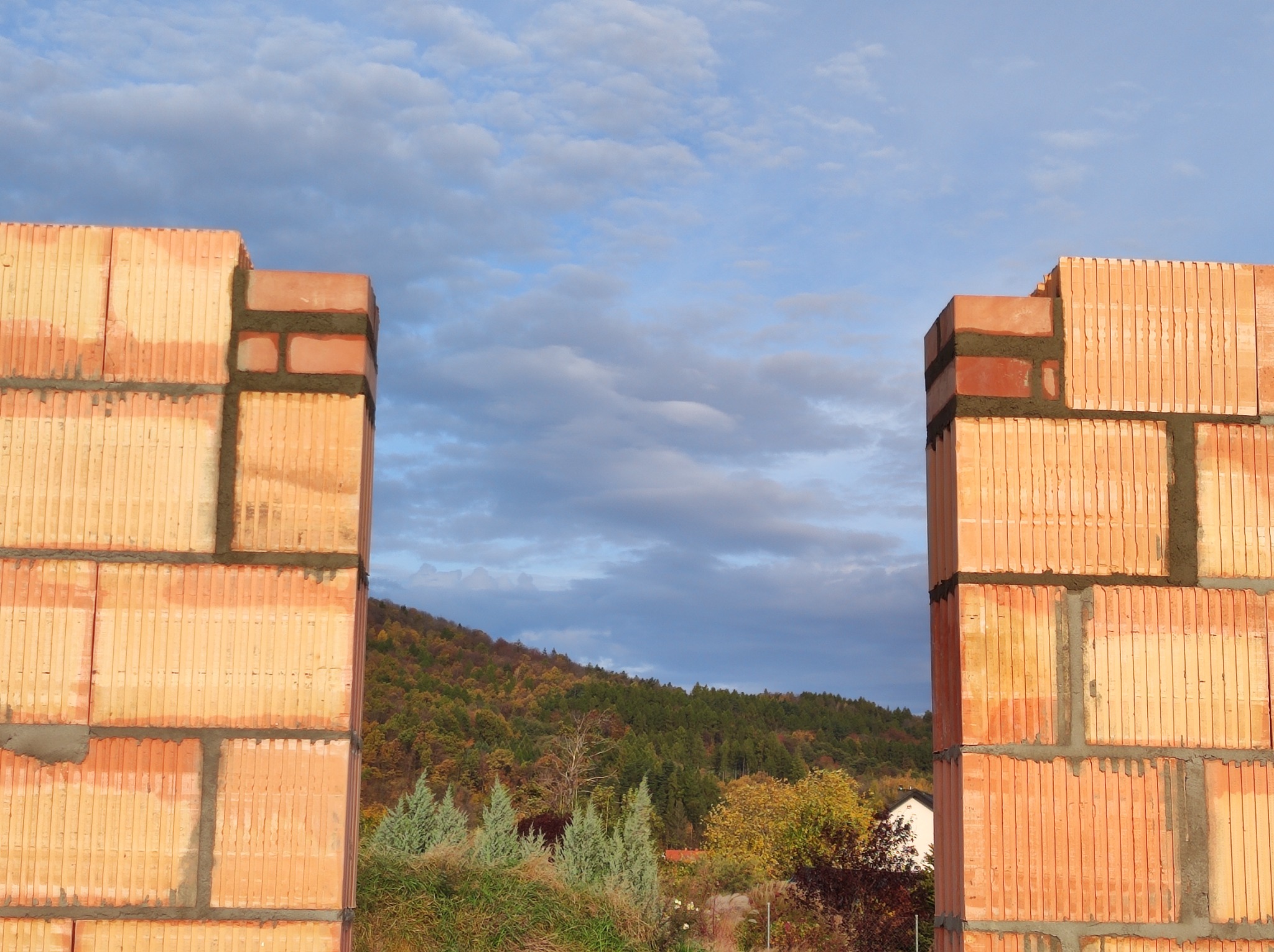 Fragment wznoszonej ściany z bloczków ceramicznych, widok na zielony pagórek i pochmurne niebo w tle.