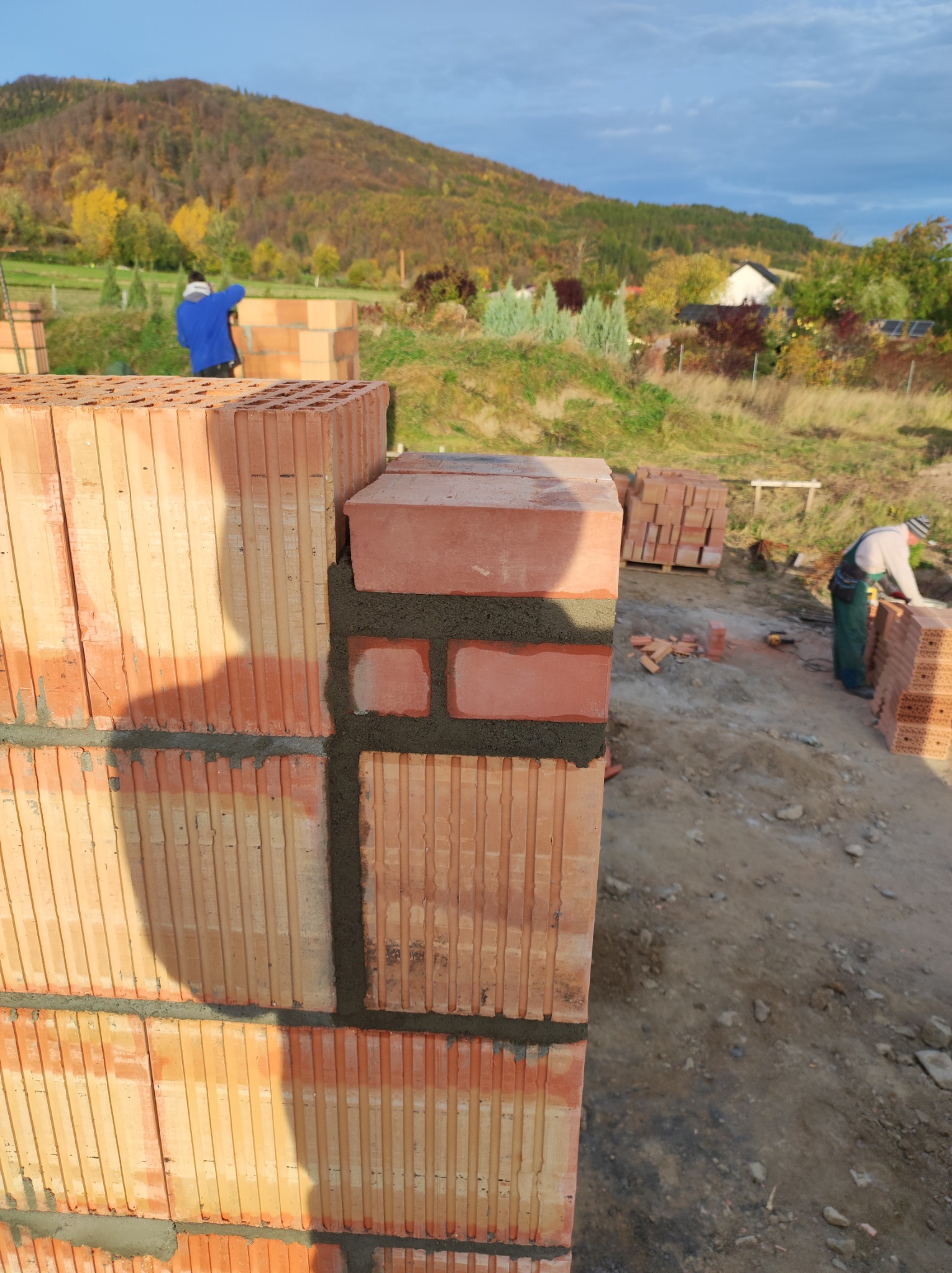 Wznoszenie ściany z bloczków ceramicznych, widoczne świeże spoiny, w tle krajobraz zalesionych wzgórz i pracujący murarze.
