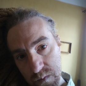 Portret mężczyzny z dredami, skierowany w lewo, z delikatnym uśmiechem, na tle ściany w odcieniach beżu i żółci, z widoczną ramką obrazu w tle.