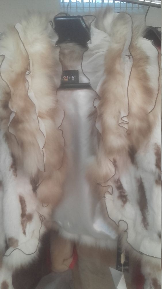 Białe futro z brązowymi plamami i falbankami, wiszące na wieszaku, widoczne metki.